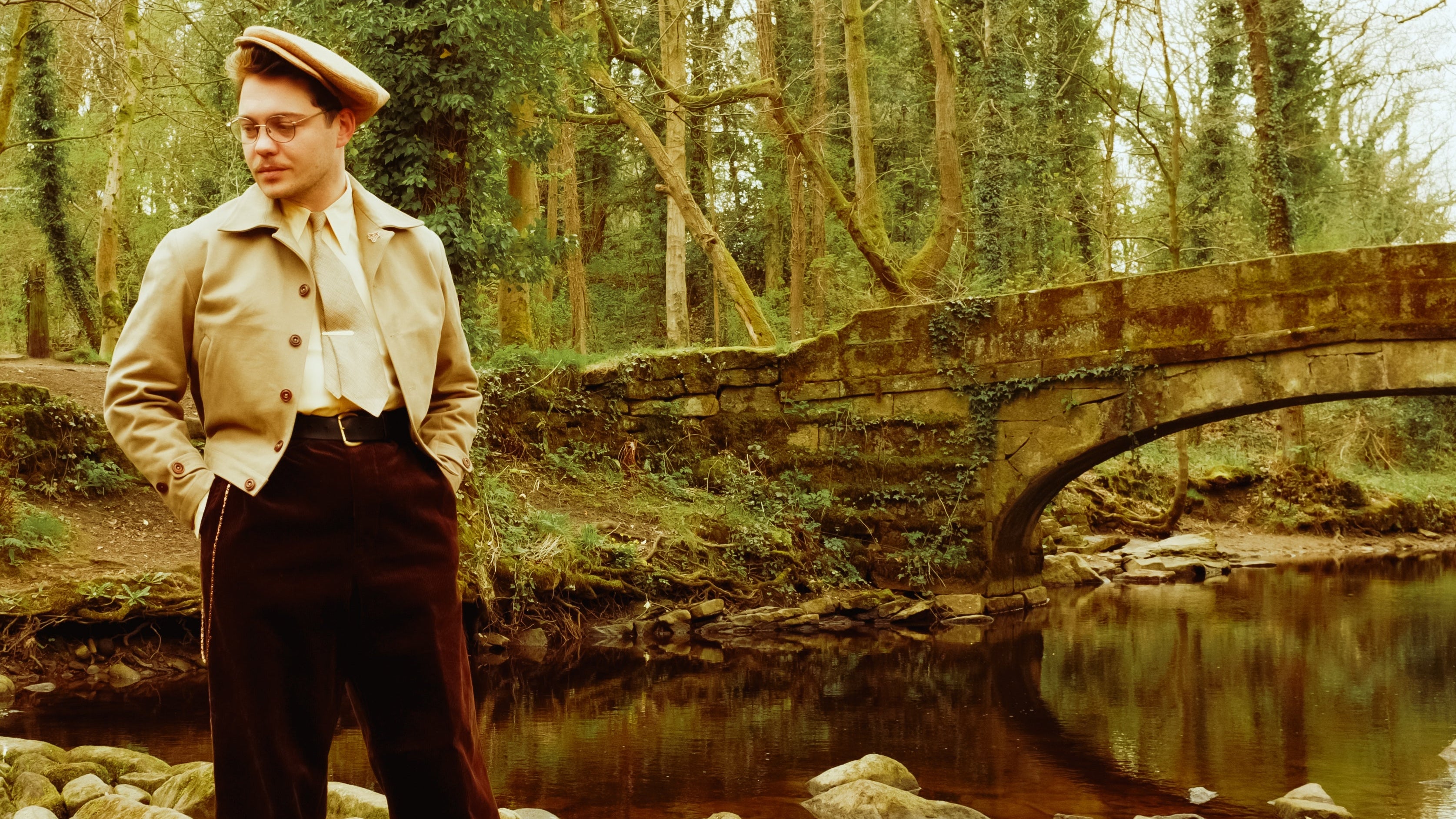 Man in vintage attire standing by a bridge over a stream in a forest