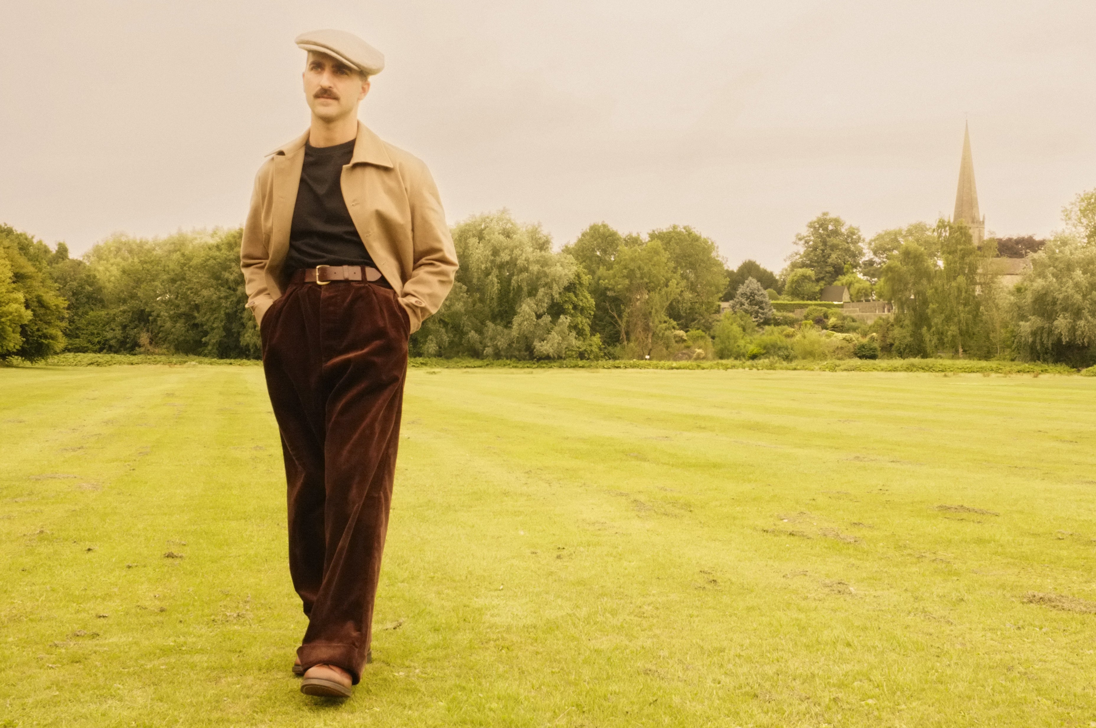 Man in vintage attire walking on a grassy field with trees and a church spire in the background