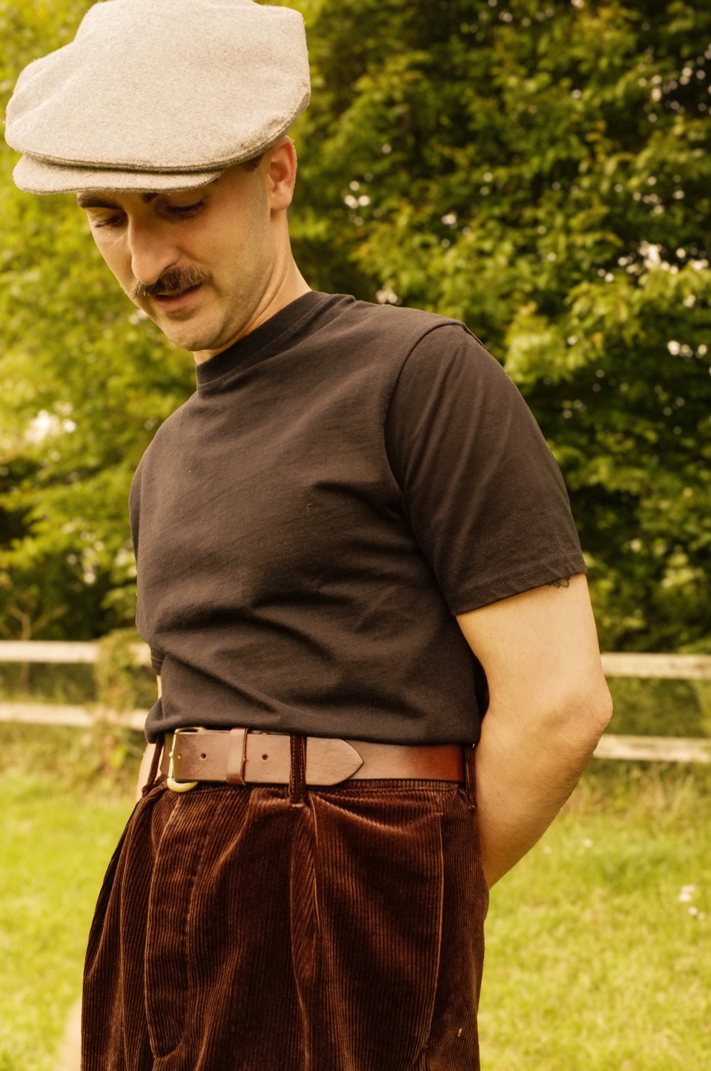 Man wearing a brown shirt, cap, and brown pants standing outdoors with greenery in the background