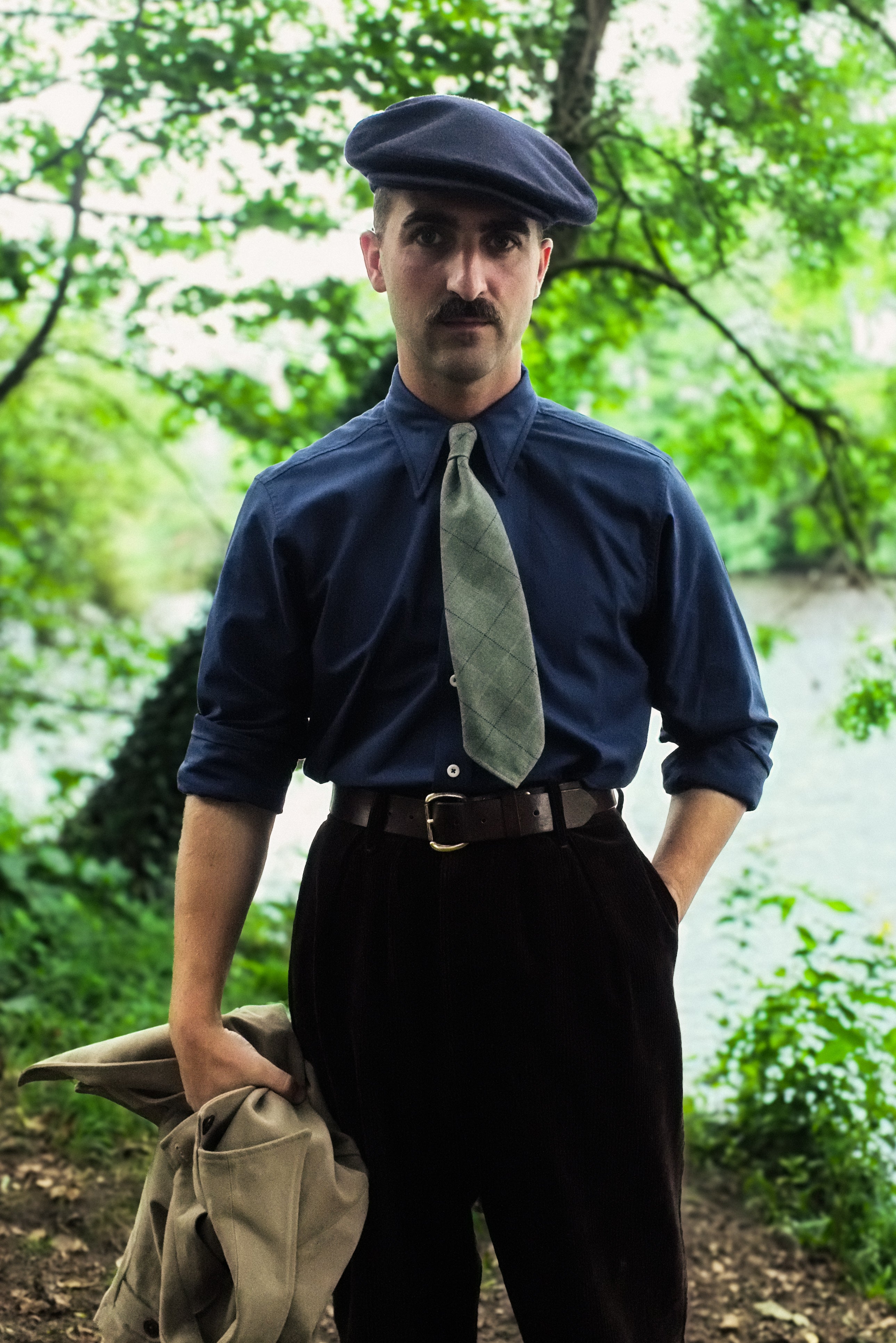 Man in vintage attire with a blue shirt, tie, and cap standing outdoors in a natural setting.